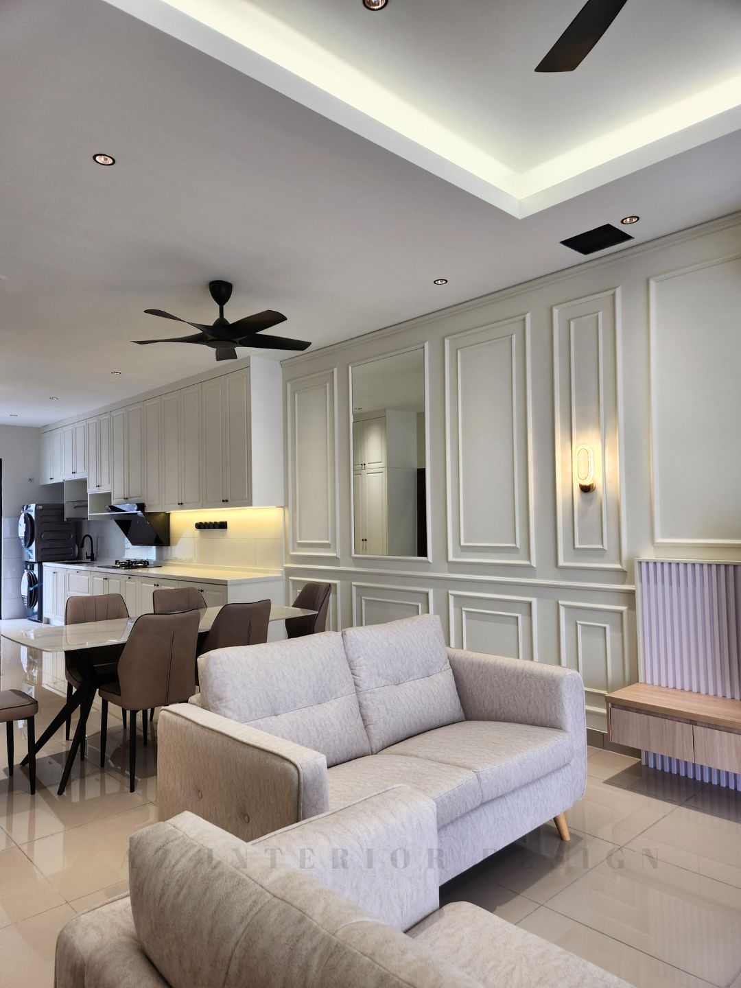 Open-plan living, dining, and kitchen area with classic European wall paneling, neutral-toned furniture, and modern ceiling fans by AZ Interior Design.