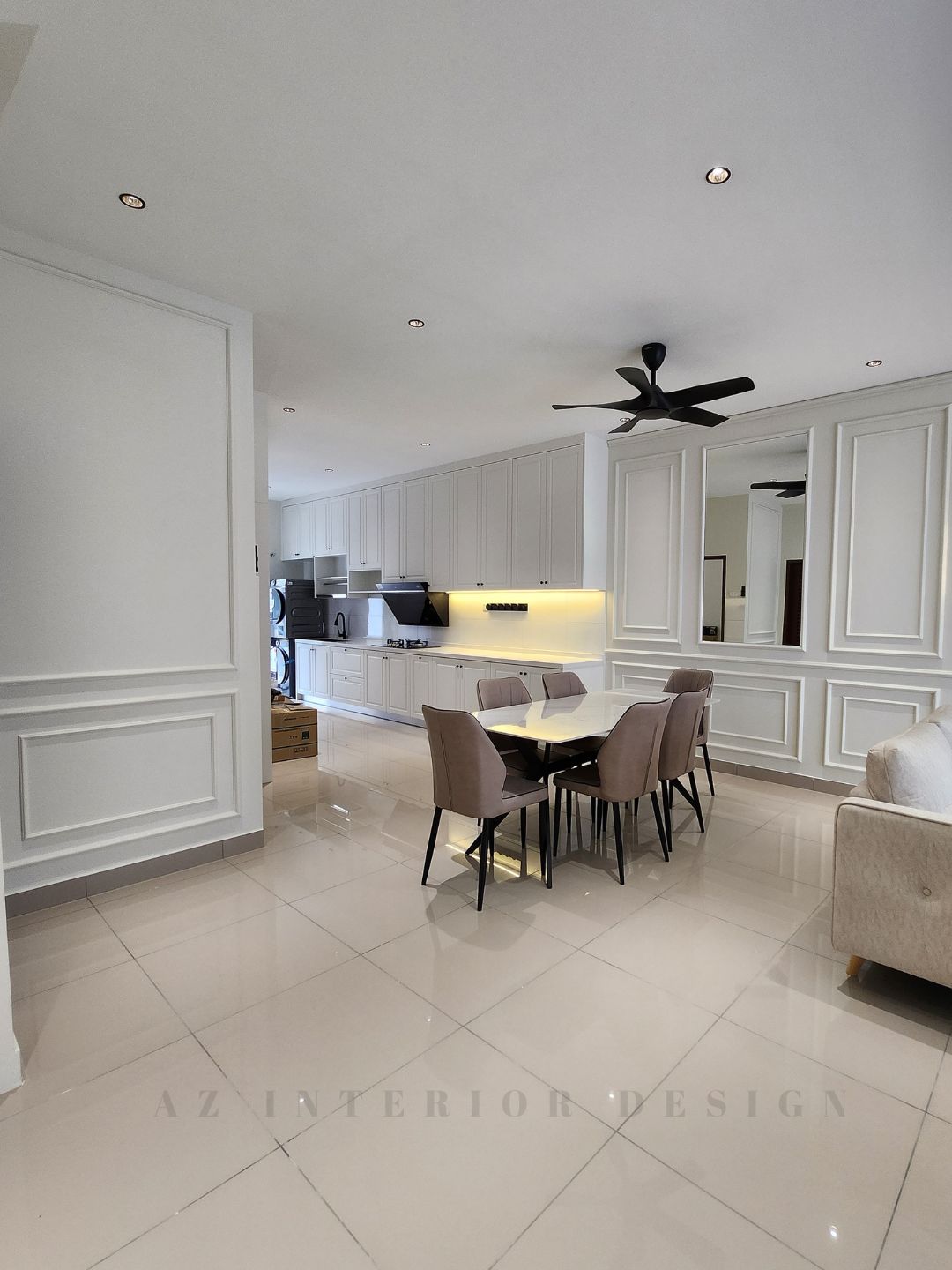 Open-plan living, dining, and kitchen area with classic European wall paneling, neutral-toned furniture, and modern ceiling fans by AZ Interior Design.