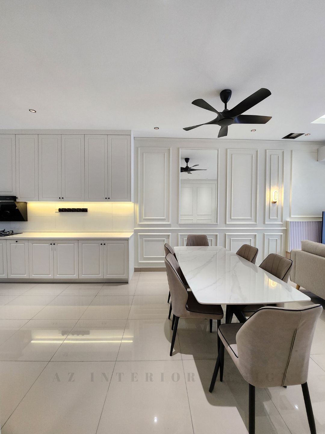 Open-plan living, dining, and kitchen area with classic European wall paneling, neutral-toned furniture, and modern ceiling fans by AZ Interior Design.